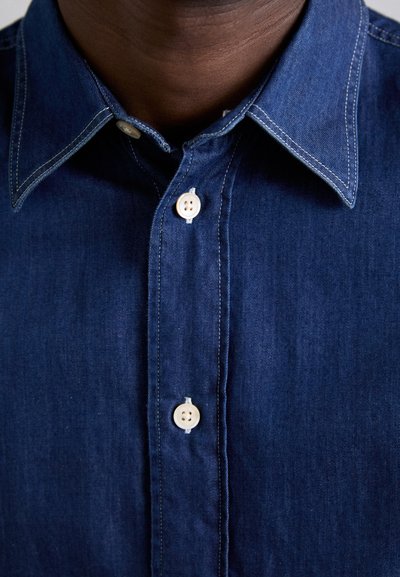 Camicia in denim scuro con colletto classico, caratterizzata da cuciture bianche e bottoni beige. Vista ravvicinata della patta dei bottoni e del colletto.