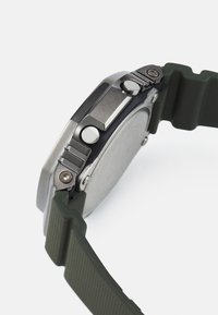 Stainless steel watch with a green rubber strap, featuring textured accents, two buttons, and a brushed metal finish on the case.