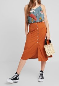Jupe midi orange avec des boutons sur le devant, associée à un débardeur fleuri et des baskets montantes noires. Comprend un sac en osier beige.