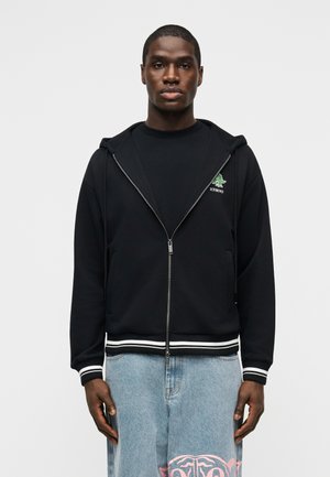 Nuori mies, joka pukeutunut mustaan vetoketjuhuppariin, jossa on vihreä logo, ja vaaleansiniset farkut, joissa on vaaleanpunaisia kuvioita, seisoo yksiväristä valkoista taustaa vasten.