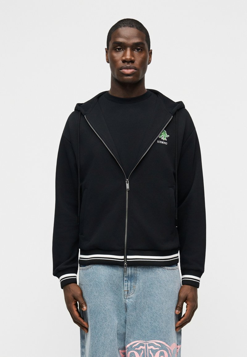 Νέος άνδρας με μαύρο φούτερ με φερμουάρ και πράσινο λογότυπο, και γαλάζια τζιν με ροζ σχέδια, στέκεται μπροστά σε λευκό φόντο.