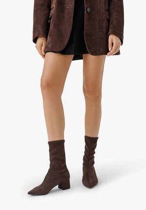 Femme portant des bottines en daim marron foncé à talons blocs, une veste marron et une jupe courte noire, debout sur un fond blanc.