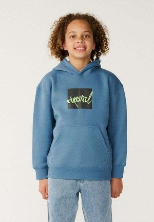 Niño con cabello rizado que lleva una sudadera azul con capucha con un bolsillo delantero y un logo cuadrado negro, de pie frente a un fondo claro y liso.