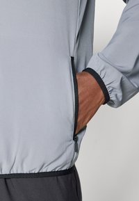 Jachetă ușoară de culoare gri deschis, cu manșete elastice și un buzunar cu fermoar, având o căptușeală interioară maro în contrast vizibilă la deschiderea buzunarului.