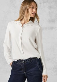 Blouse blanche à manches longues avec un devant boutonné et un col classique, associée à un jean en denim bleu foncé avec des coutures contrastantes.