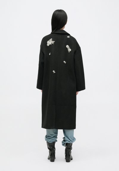 Črn plašč z strukturirano obliko, okrašen s srebrnimi detajli, razporejenimi po hrbtu, v kombinaciji z svetlo modrimi kavbojkami in črnimi škornji.