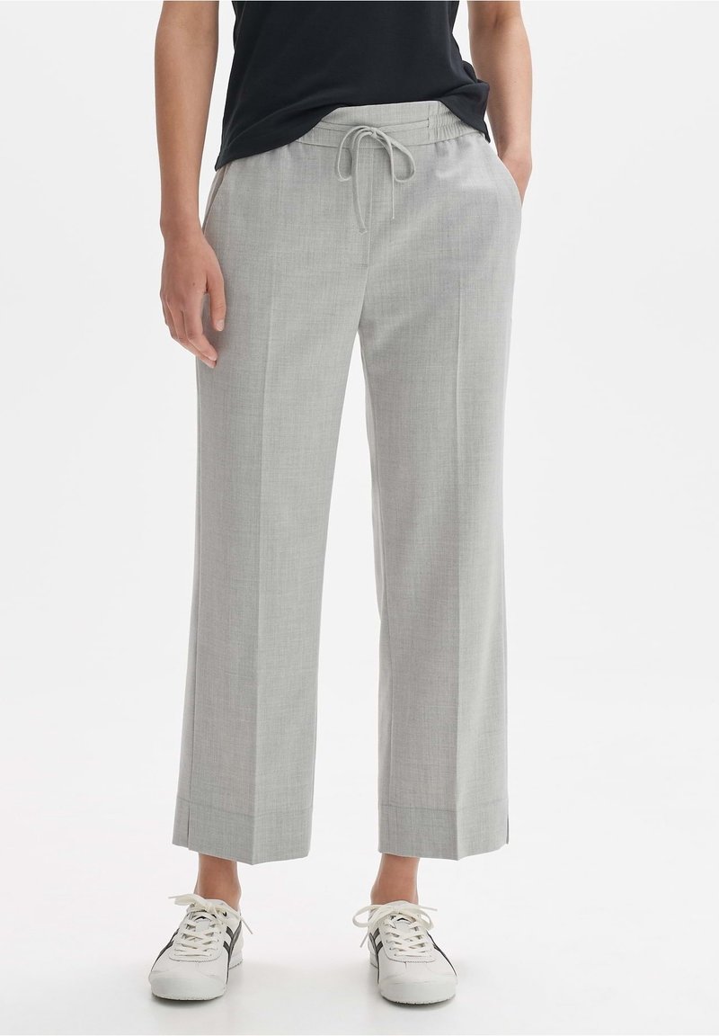 Personne portant un pantalon large gris clair avec taille à cordon, un haut noir et des baskets blanches avec des rayures noires, debout sur fond blanc.