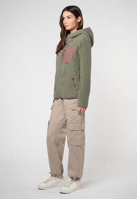 Fleecejacke in Olivgrün mit einer braunen Brusttasche, Reißverschluss vorne und Kapuze; kombiniert mit beigen Cargohosen und weißen Sneakern.
