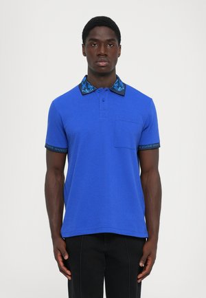 Male wearing a bright blue polo shirt with patterned collar and black pants, standing facing forward against a plain light background.