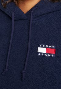Felpa con cappuccio in pile blu navy, dotata di tasca a marsupio, cordini regolabili e una patch rettangolare con la scritta "TOMMY JEANS" in bianco e rosso.