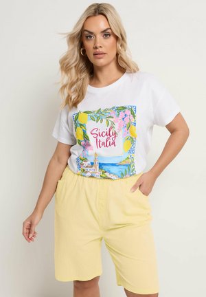 Blonde Frau trägt weißes T-Shirt mit buntem Sicily Italia-Aufdruck und gelbe Shorts, posiert mit einer Hand in der Tasche vor schlichtem Hintergrund.