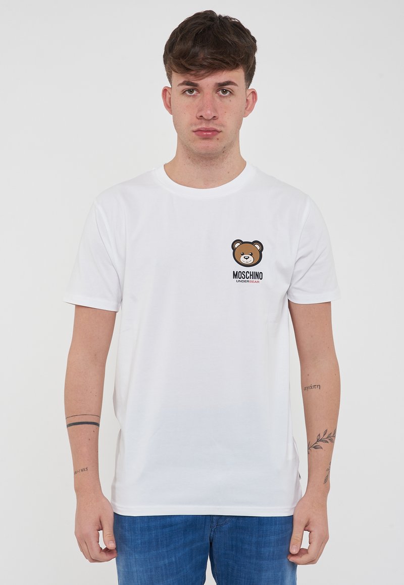 T-shirt in cotone bianco con grafica di un orso bruno sul lato sinistro del petto e testo "Moschino Underwear" in nero sottostante. Maniche corte.