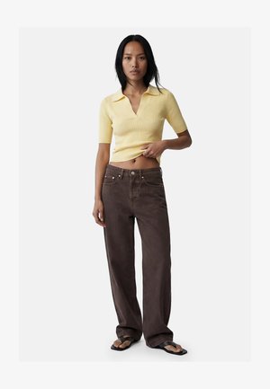 Frau mit langen schwarzen Haaren, die ein gelbes kurzärmeliges Oberteil mit Kragen, braune weite Jeans und schwarze Flip-Flop-Sandalen trägt, steht vor weißem Hintergrund.