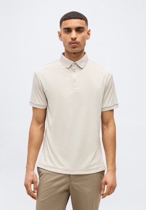 Νέος άνδρας με κοντά μαλλιά που φοράει μπεζ κοντομάνικο πόλο πουκάμισο και ανοιχτό καφέ παντελόνι, στέκεται μπροστά σε απλό φωτεινό φόντο.