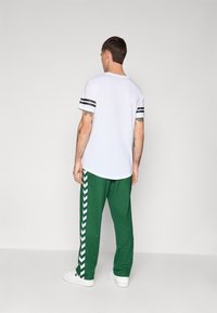 Férfi hátulról áll, fehér pólót visel fekete csíkokkal az ujjakon, zöld melegítőnadrágot fehér chevron mintákkal és fehér tornacipőt.