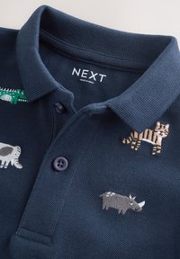 Polo bleu foncé avec des animaux brodés, dont un tigre, un rhinocéros, un éléphant et un crocodile vert près du col.