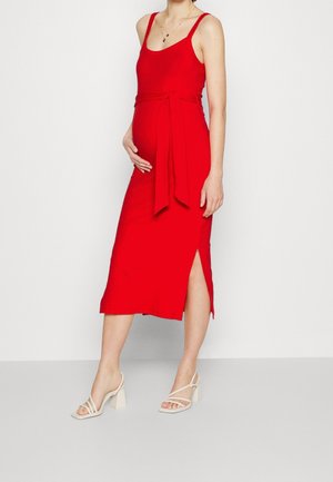 Mujer embarazada con vestido midi rojo sin mangas con abertura lateral y sandalias beige de tacón, de pie frente a un fondo claro y liso.