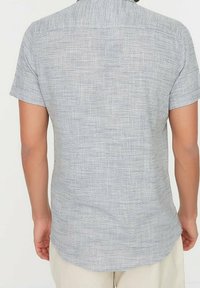 Camisa de manga corta en tejido gris claro con un patrón texturizado. Presenta un pequeño cuello y un dobladillo curvado, vista trasera.