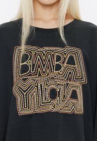 Felpa nera con testo ricamato multicolore "BIMBA" e "YLOLA," caratterizzata da cuciture spesse e testurizzate in rosa, giallo e nero.