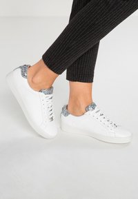 Sneakers in pelle bianca con accenti di glitter argento sul tallone, caratterizzati da punta rotonda, design con lacci e una robusta suola in gomma bianca.
