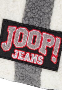 Teksturowany materiał w paski w odcieniach szarości i kremu. Zawiera czarną łatę z napisem "JOOP!" w czerwonym i szarym, oraz "JEANS" w białym, wyraźnie wyszytym.