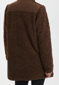 Hnědá chlupatá fleecová bunda s hladkou hnědou horní částí. Volný střih, s kulatým lemem a dlouhými rukávy. Bez viditelných zapínání.