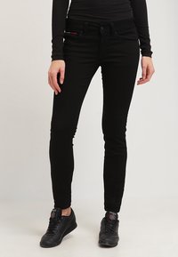 Jeans ajustados negros de denim, con cremallera en la bragueta, bolsillos frontales y una textura suave y lisa. Llevados con zapatillas deportivas negras.