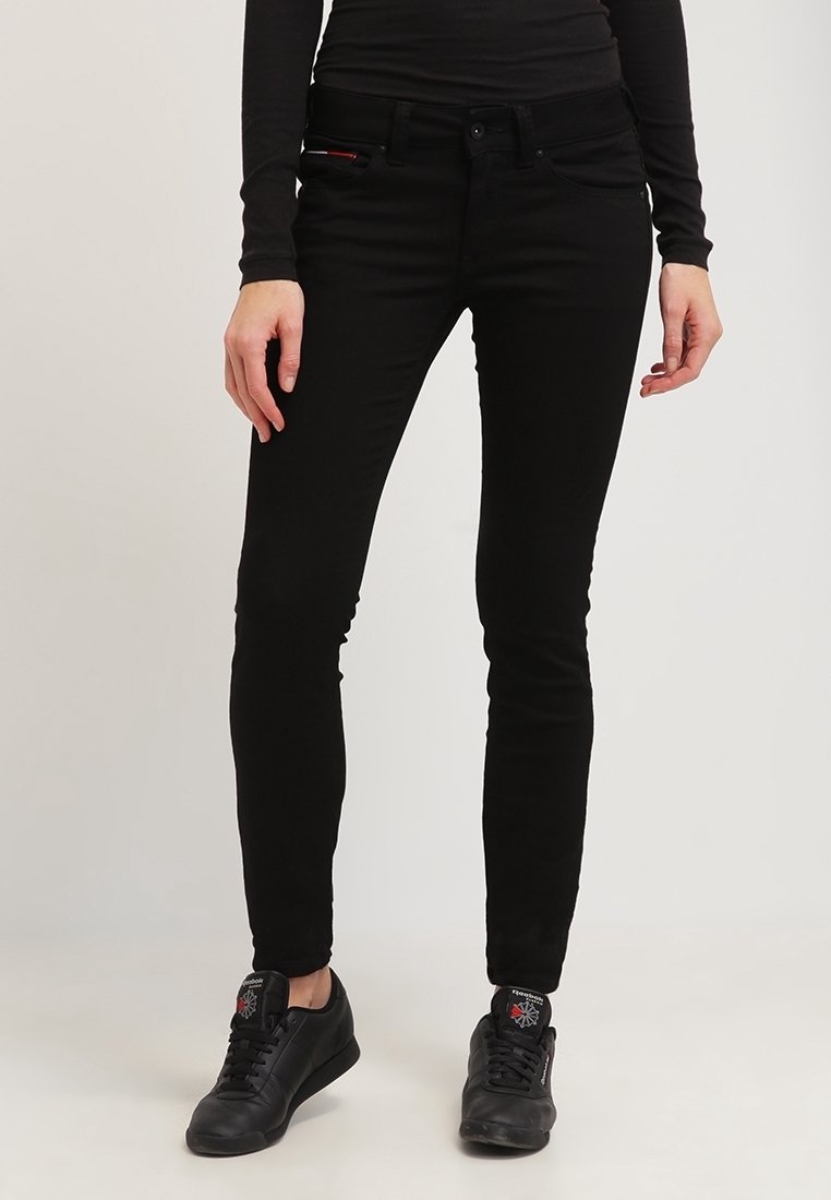 Jeans ajustados negros de denim, con cremallera en la bragueta, bolsillos frontales y una textura suave y lisa. Llevados con zapatillas deportivas negras.