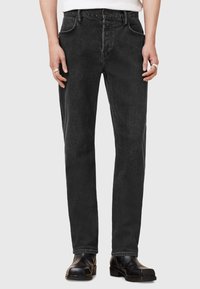 Jeans de mezclilla negra con corte recto, cinco bolsillos y un desgaste sutil, combinados con calzado negro. Cintura estándar y cierre de cremallera.