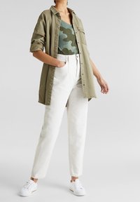 Camisa de botón verde oliva sobre una camiseta de camuflaje, combinada con pantalones blancos de tiro alto y zapatillas blancas. Un look suave y casual.