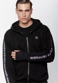 Czarna bluza z kapturem na zamek, uszyta z miękkiego materiału, z ribbowanymi mankietami i naszywką z logo. Rękawy po bokach mają powtarzający się graficzny wzór w kolorze białym.