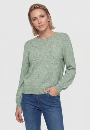 Donna con capelli biondi corti che indossa un maglione verde chiaro lavorato a maglia e jeans blu, in piedi con una mano in tasca davanti a uno sfondo uniforme.