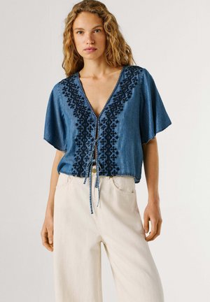 Vrouw draagt een blauw geborduurd, kort mouw blouse met strikjes aan de voorkant en een hoog taillieuze, wijde crème broek, staand tegen een effen achtergrond.