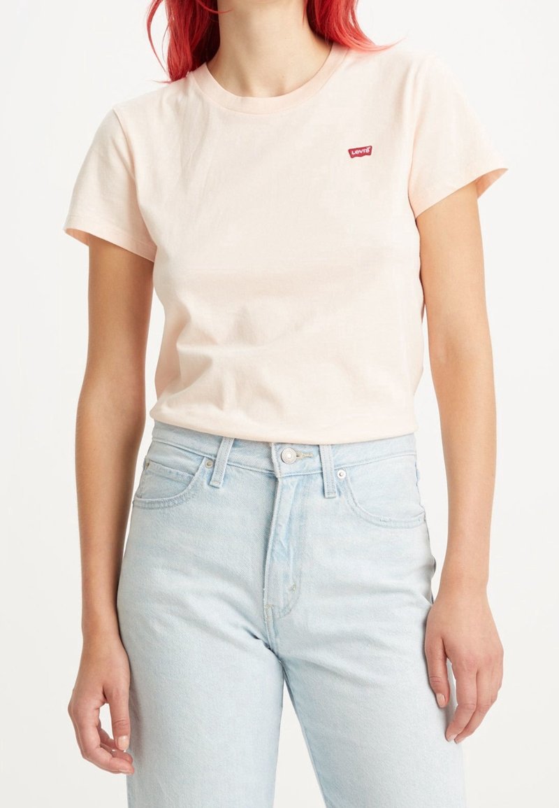 Personne portant un t-shirt Levi's rose pâle et un jean taille haute bleu clair, debout devant un fond blanc uni.