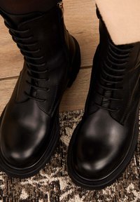 Stivaletti neri in pelle con una texture liscia, design con lacci, suola spessa e dettaglio con zip laterale, posizionati su una superficie decorata.