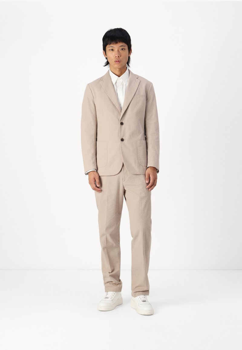Light beige suit made of textured material with a single-breasted jacket, two pockets, and matching trousers, paired with white sneakers.