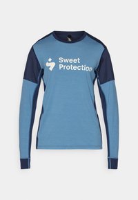 Majica dugih rukava plave boje s mornarsko plavim detaljima. Ima okrugli izrez i bijeli logo "Sweet Protection" sprijeda. Glatka tekstura.