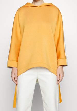 Personne portant un sweat à capuche jaune ample à manches trois-quarts et un pantalon blanc, debout devant un fond clair uni.