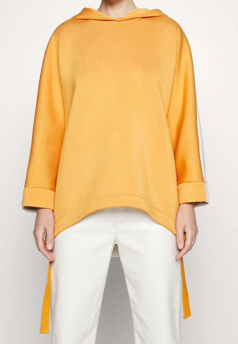 Personne portant un sweat à capuche jaune ample à manches trois-quarts et un pantalon blanc, debout devant un fond clair uni.