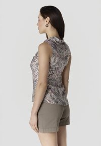 Mujer con cabello largo y oscuro, vestida con una blusa sin mangas de estampado abstracto y pantalones cortos beige, de pie de perfil contra un fondo liso.