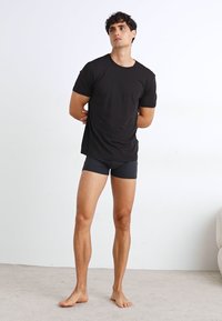 T-shirt noir à col ras du cou associé à des boxers gris foncé. Modèle debout sur un tapis clair, pieds nus, les bras croisés derrière le dos.