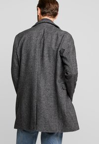 Abrigo gris de lana con patrón de espiga, cuello con muesca y dos bolsillos laterales, combinado con vaqueros azul claro.