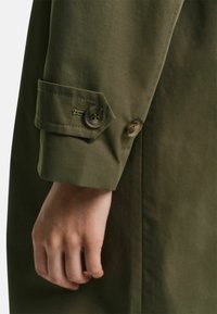 Ruka v olivově zeleném rukávu s dvěma hnědými knoflíky na manžetě, látka se zdá být hladká a matná.