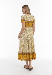 Virágmintás maxi ruha rövid ujjakkal, fehér alap, sárga díszítésekkel és réteges szegéllyel. Jellemzője a mintás bodice és az áramló szoknya.