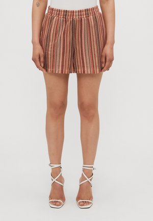 Femme portant un short rayé vertical multicolore avec une taille élastique et des sandales blanches à talons hauts avec des brides, debout devant un fond uni.