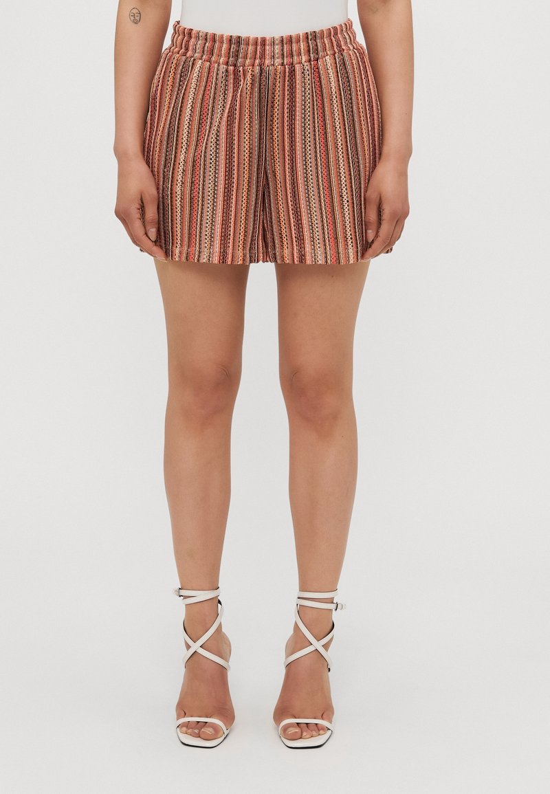 Femme portant un short rayé vertical multicolore avec une taille élastique et des sandales blanches à talons hauts avec des brides, debout devant un fond uni.