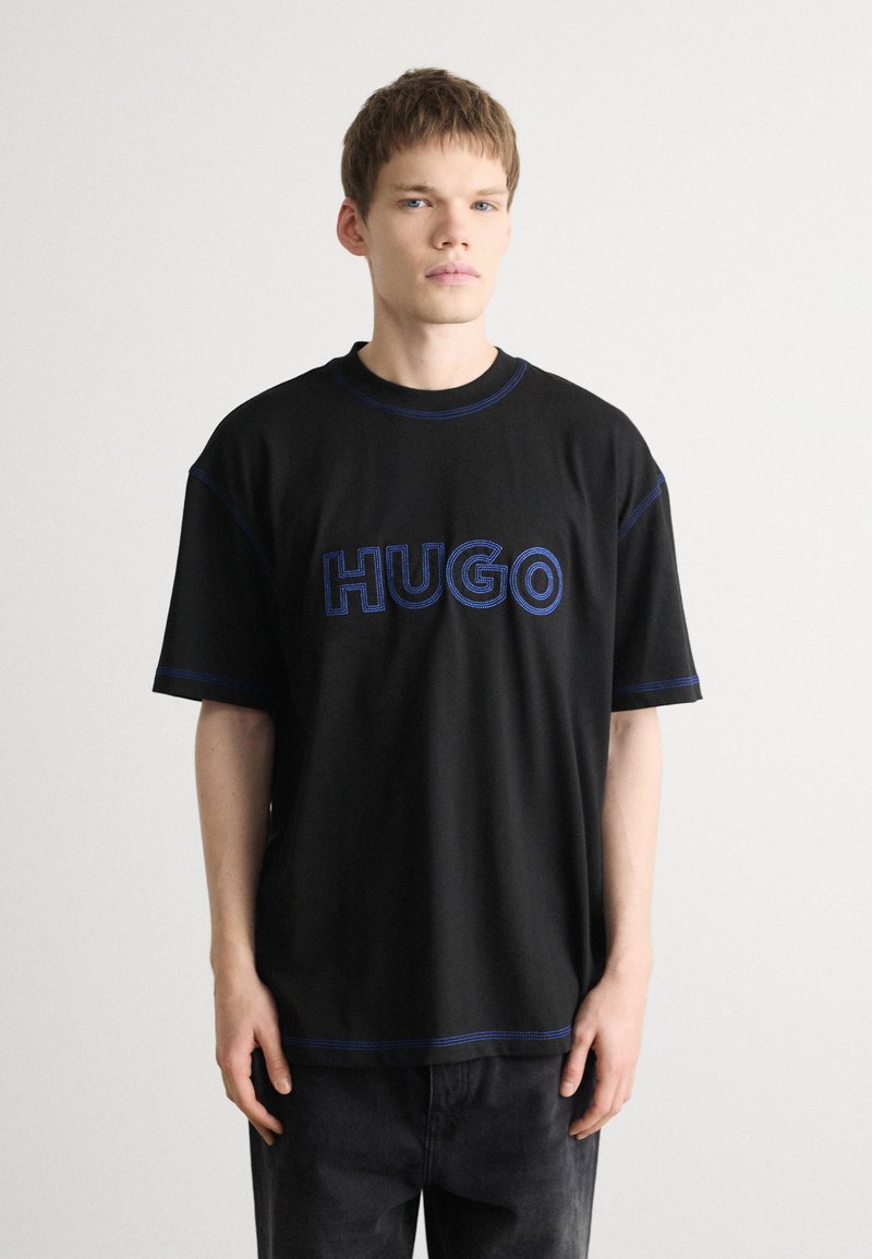 Melna īsām piedurknēm T-krekls ar zilu "HUGO" logotipa izšuvumu un kontrastējošiem ziliem vīlēm ap kakla izgriezumu un apakšu.