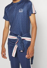 Granatowa koszulka sportowa z geometrycznym wzorem, krótki rękaw i haftowane logo; połączona z dopasowanymi spodniami z kontrastowymi paskami.