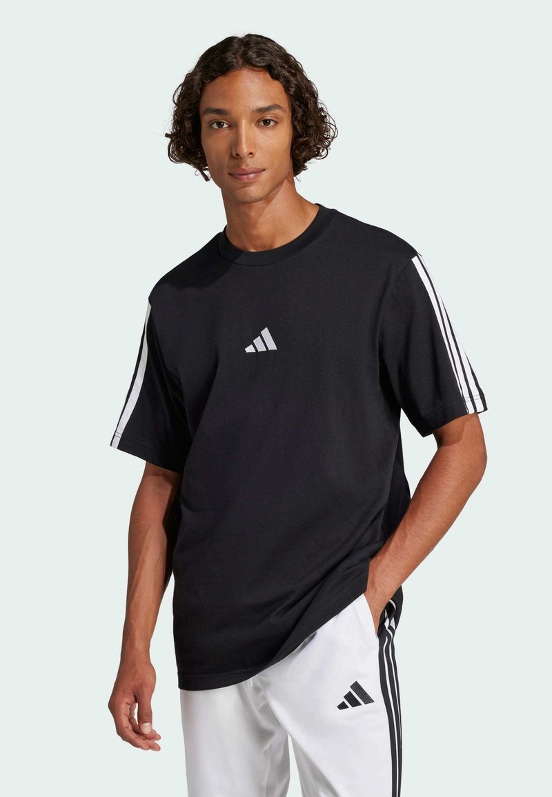 Černé tričko s krátkým rukávem a bílým logem Adidas, s třemi bílými pruhy na každém rameni, vyrobené z hladké látky a s volným střihem.