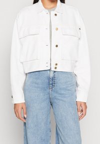 Giacca cropped in denim bianco con due tasche frontali, bottoni dorati e colletto strutturato, abbinata a jeans a gamba larga azzurri.